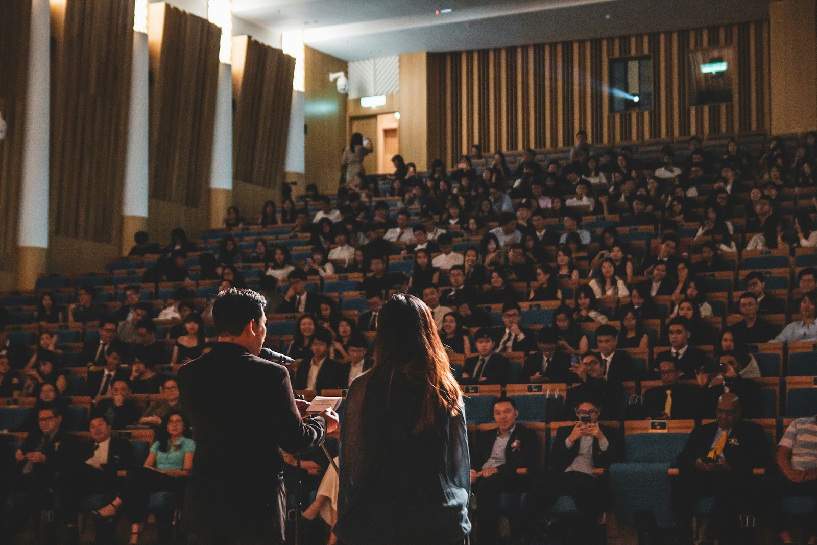 An image of a man and a woman holding a speech in front of the audience.